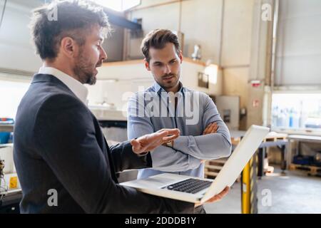 Mature man using laptop while standing by colleague at factory Stock Photo