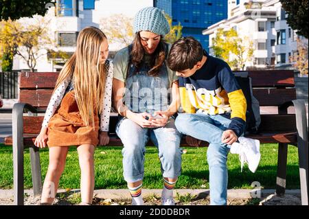 Mother with kids using smart phone sitting on bench in public park during sunny day Stock Photo