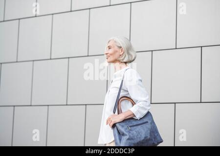 Smiling businesswoman with purse looking away while standing against wall Stock Photo