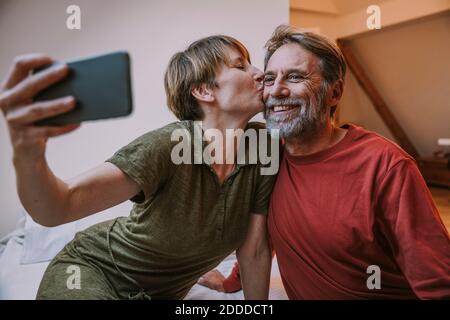 Woman kissing mature man while taking selfie on smart phone sitting in bedroom at home Stock Photo