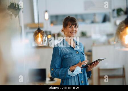 Female entrepreneur using digital tablet while holding protective face mask standing in cafe Stock Photo