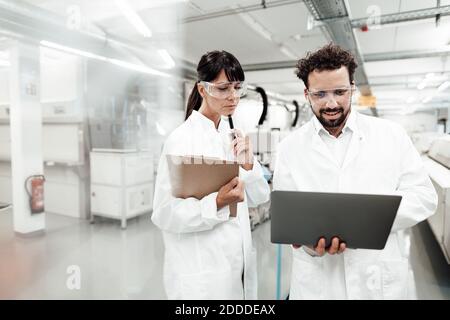 Smiling male scientist using laptop standing by female technician in laboratory Stock Photo