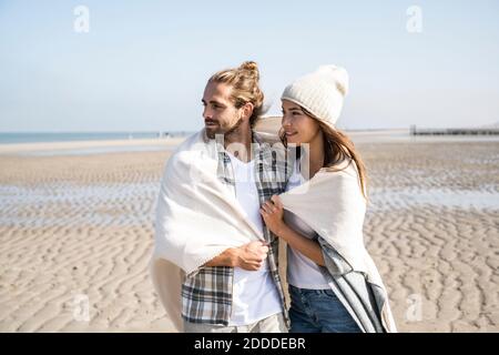 Thoughtful young couple covered in blanket looking away while standing at beach on sunny day Stock Photo