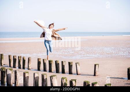 Cheerful woman with arms outstretched walking on wooden posts at beach during sunny day Stock Photo