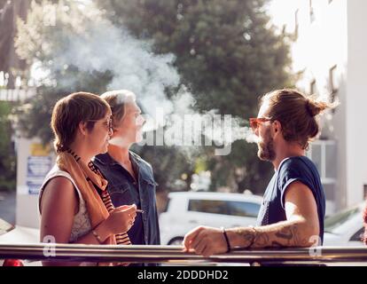 Friends smoking while standing outdoors on sunny day Stock Photo