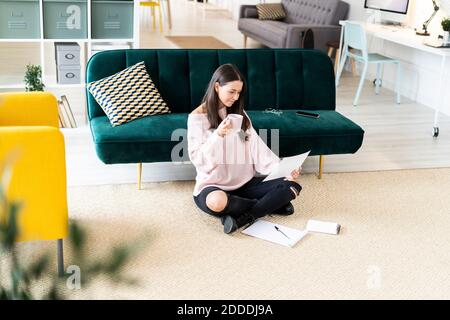 Beautiful young woman drinking coffee while reading paper by note pads on carpet against sofa at home Stock Photo