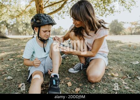 Mother putting bandage on son's knee while sitting in public park during sunny day Stock Photo