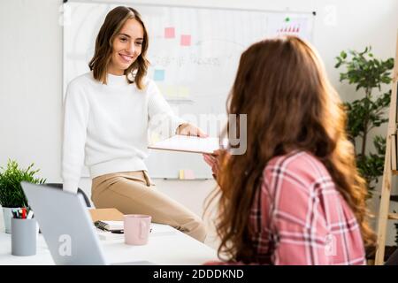 Smiling businesswoman giving notepad to colleague while sitting on tablet at office Stock Photo