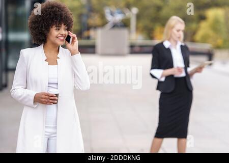 Businesswoman talking on mobile phone while standing with colleague in background on footpath Stock Photo