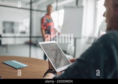 Colleague looking at diagram on digital tablet screen while sitting with businessman in background at office Stock Photo
