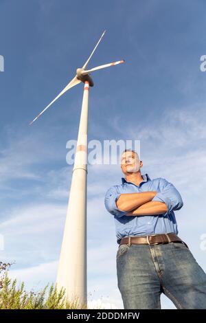 Confident mature male engineer standing with arms crossed against wind turbines on field Stock Photo