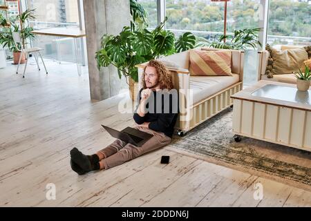 Man with hand on chin using laptop sitting in living room at home Stock Photo