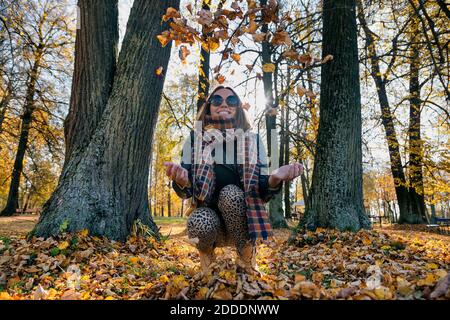 Cheerful woman throwing dry autumn leaves while crouching against trees at park Stock Photo