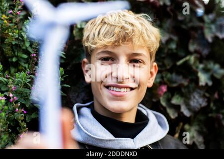 Smiling blond teenage boy holding wind turbine model against plants Stock Photo