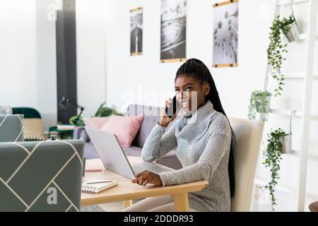 Smiling female student talking on smart phone while studying through laptop at home Stock Photo