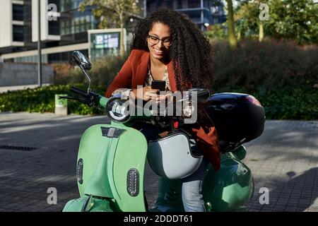 Smiling woman using smart phone while sitting on motor scooter during sunny day Stock Photo