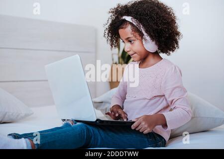 Cute girl using laptop wearing headphone listening music while sitting in bedroom at home Stock Photo