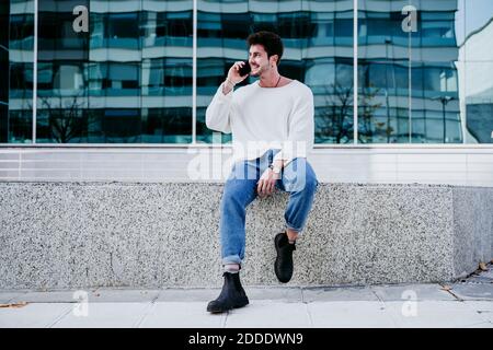 Smiling man talking on smart phone while sitting on retaining wall against building in city Stock Photo