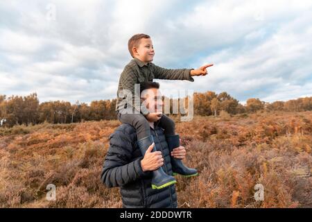 Father carrying son on shoulder standing in Cannock Chase park against cloudy sky during autumn Stock Photo