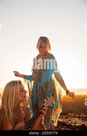 Young woman playing ukulele while female friend dancing at beach during sunset Stock Photo