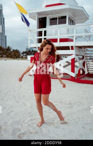 Young woman looking down while playing on sand against lifeguard house at beach Stock Photo