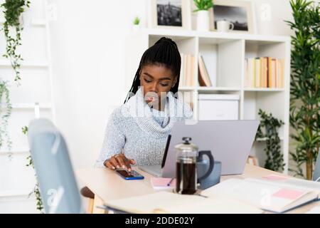 Young female student using smart phone while studying through laptop sitting in living room at home Stock Photo