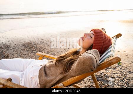 Young woman relaxing on folding chair with eyes closed at beach during sunset