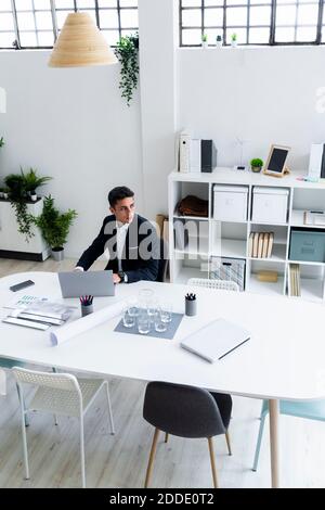 Thoughtful young male entrepreneur looking away while sitting with laptop at desk in workplace Stock Photo