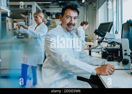 Smiling male scientist sitting by microscope while coworker working in background at laboratory