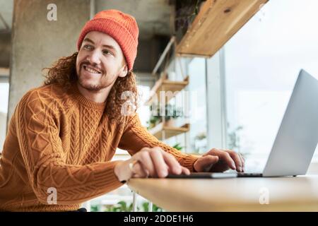 Smiling hipster man looking away while using laptop at home Stock Photo