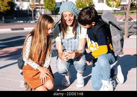 Mother using smart phone crouching with kids in public park on sunny day Stock Photo