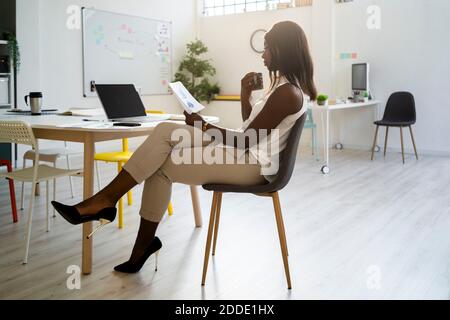 Woman with coffee cup reading paper while sitting on chair at office Stock Photo