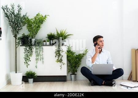 Businessman talking through mobile phone while sitting with laptop on floor against white wall in office Stock Photo