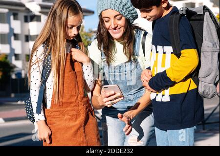 Mother using smart phone standing with kids in public park on sunny day Stock Photo
