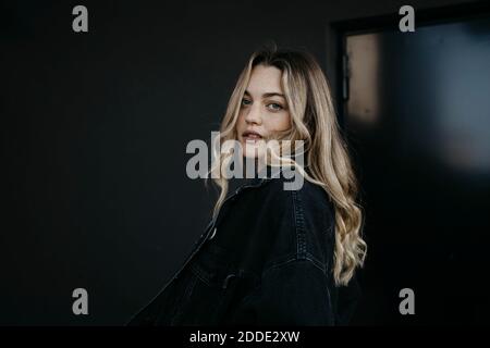 Young woman wearing denim jacket while standing against black wall Stock Photo