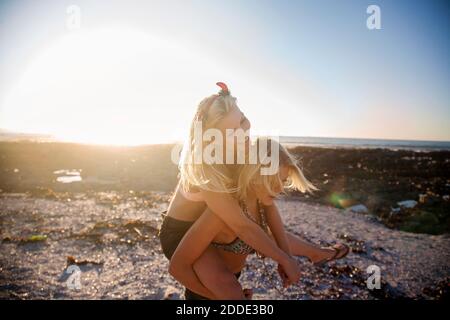 Young woman piggybacking cheerful female friend at beach during sunset Stock Photo