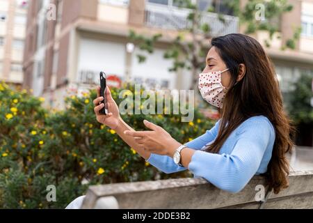 Woman gesturing while using smart phone on bench in city during COVID-19 Stock Photo