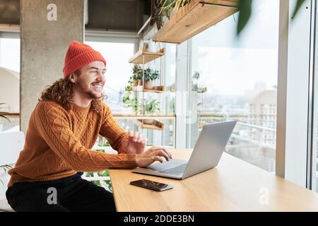Smiling man wearing knit hat using laptop sitting at home Stock Photo