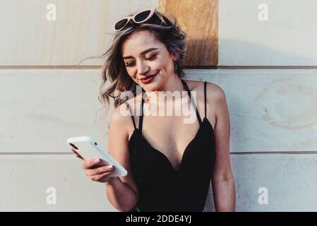Young woman text messaging on smart phone while standing against wall Stock Photo
