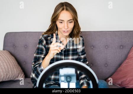 Young woman live streaming through mobile phone attached with ring flash while sitting on sofa at home Stock Photo