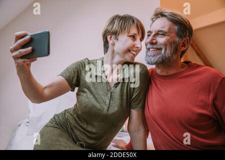 Smiling woman taking selfie while mature man sitting beside on bed at home Stock Photo