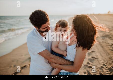 Mother and father embracing son while standing at beach during sunset