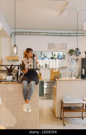 Female owner talking on smart phone sitting near counter in coffee shop Stock Photo