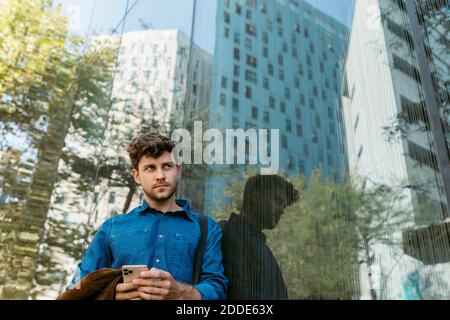 Thoughtful young businessman leaning on glass wall of modern office building Stock Photo