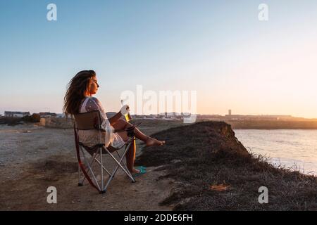 Woman drinking white wine while sitting on chair at beach during sunset Stock Photo