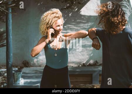 Smiling woman giving elbow bump to man while talking on mobile phone outdoors Stock Photo