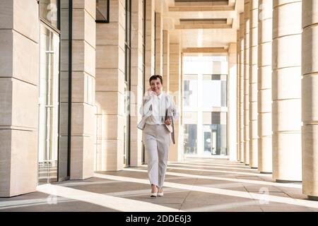Businesswoman talking on mobile phone while walking at office building corridor Stock Photo