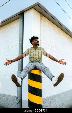 Man with arms outstretched siting on bollard against building in city Stock Photo