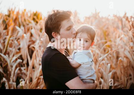Father kissing baby boy while standing against field during sunset Stock Photo