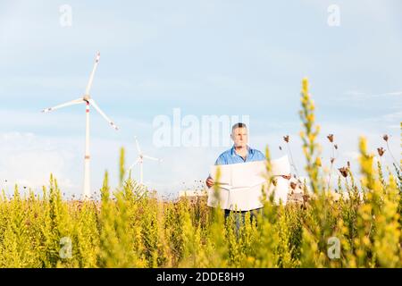 Male entrepreneur holding blueprint while standing against wind turbines on field Stock Photo